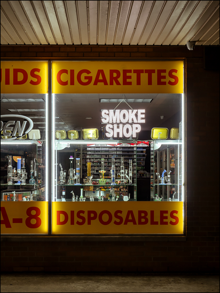 The front windows of Waynedale Smoke and Vape, a smoke shop on Bluffton Road in the Waynedale section of Fort Wayne, Indiana. The store was photographed at night. The window is filled with glass bongs that glow in the darkened window.