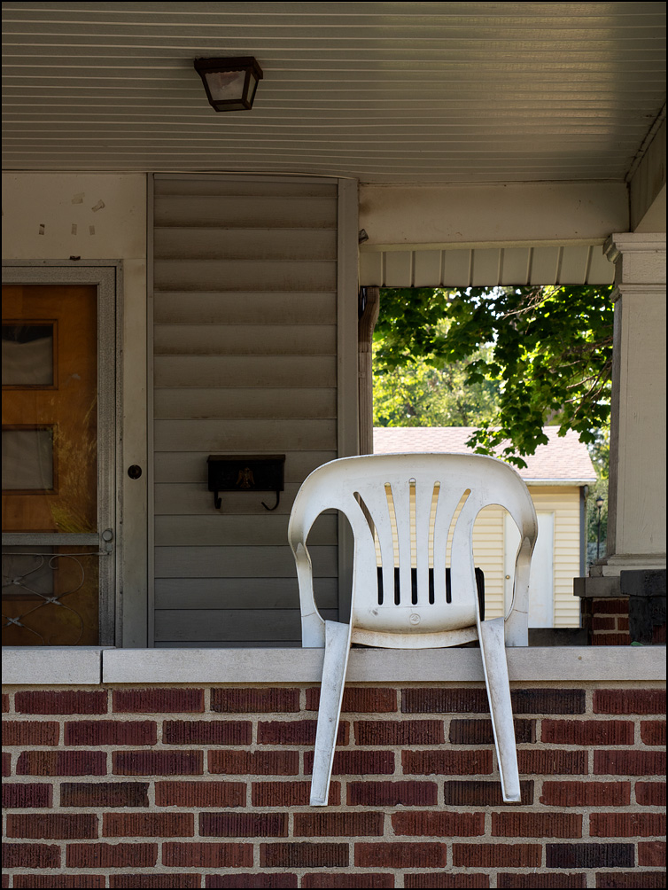 A white plastic patio chair straddling the top of the brick porch rail on a house on Cortland Avenue in Fort Wayne, Indiana.
