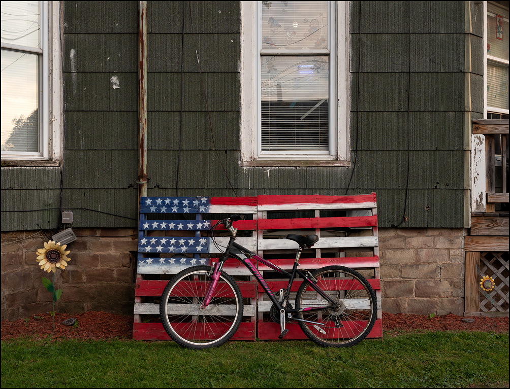 A ladies schwinn bicycle in front of a large pallet flag made of two wooden pallets next to an old green house with masonite siding on the corner of West Avenue and Eagle Street in the small town of Medina, New York.