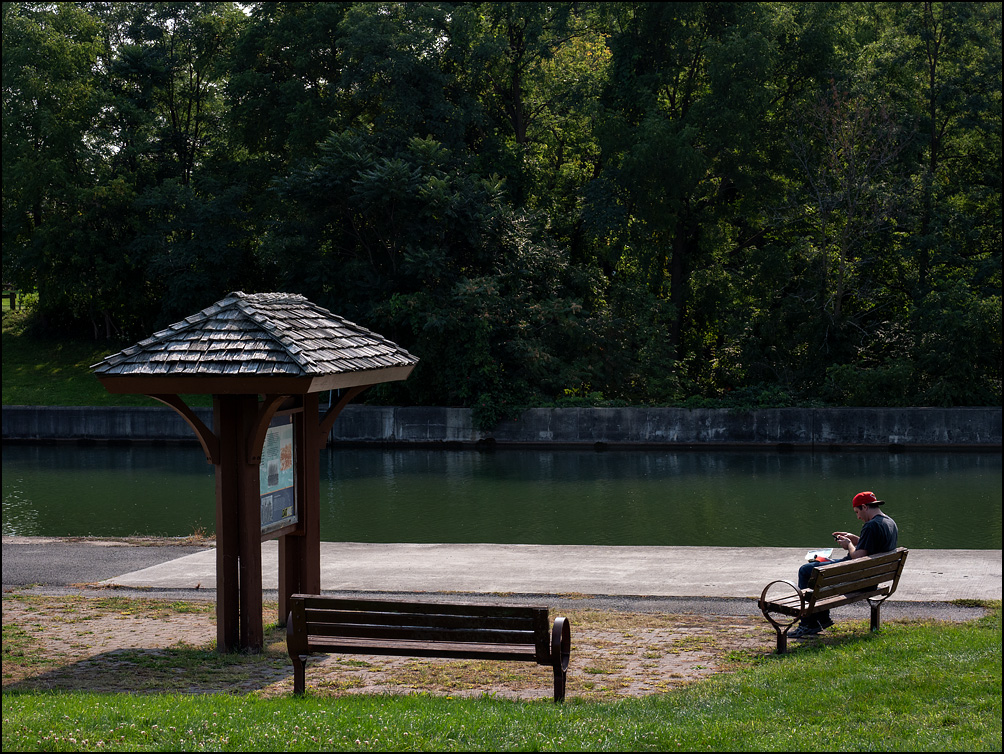 A young man sitting on a bench looking at his cellphone and eating lunch in a small park on the north bank of the Erie Canal in the small town of Medina, New York.