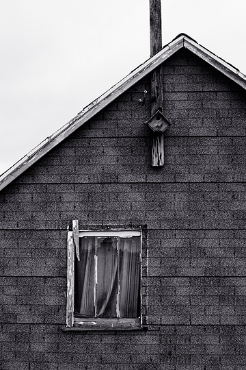 A small weathered wood birdhouse hangs on the side of an abandoned farmhouse in rural Pulaski County, Indiana. The house has asphalt shingles for siding.