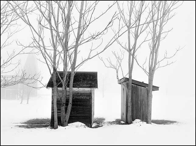 An outhouse and a small shed sit side by side among a group of trees behind the veil of a heavy winter fog on a farm in southwest Allen County, Indiana.
