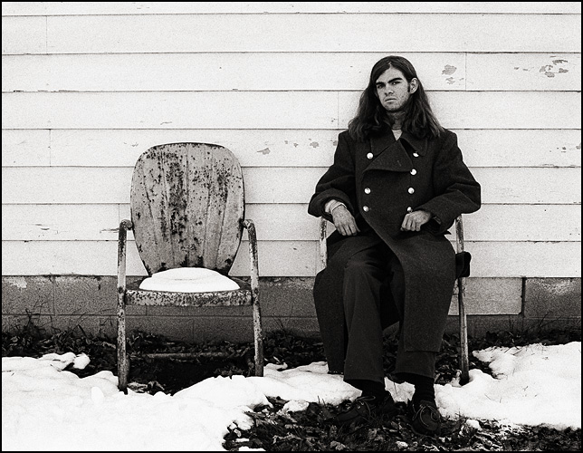 A self-portrait by fine art photographer Christopher Crawford. Chris is sitting on an old metal motel chair in the snow wearing a Soviet Army officers wool overcoat.