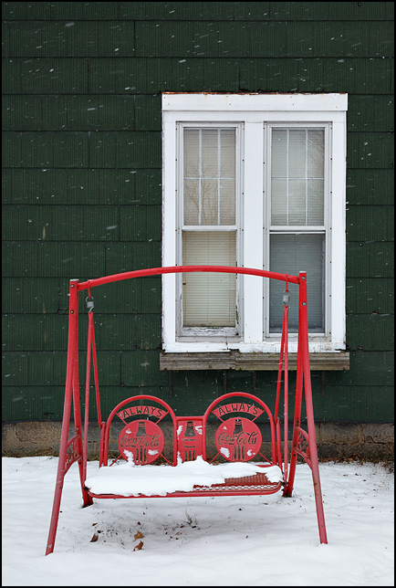 A red Coca-Cola porch swing in front of an old green house on Lincoln Highway in Goshen, Indiana. Photographed during a snowstorm.