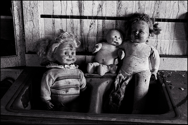 A Cricket Doll, a Cabbage Patch Kid, and a doll with one leg sit together in the kitchen sink of an abandoned house.