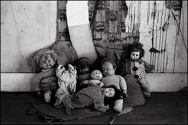Seven old broken dolls posing together against peeling wallpaper in an abandoned house.