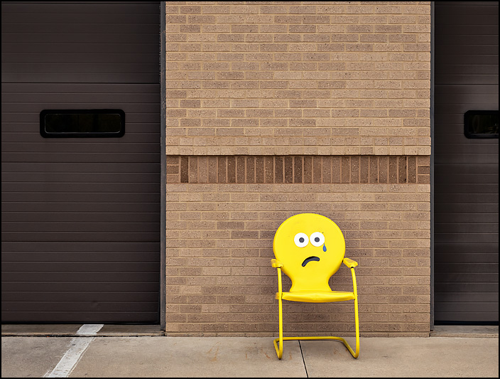 A yellow metal motel chair with a sad crying face emoji on it sits in front of Fire Station 10 at the corner of Crescent and Anthony in Fort Wayne, Indiana.