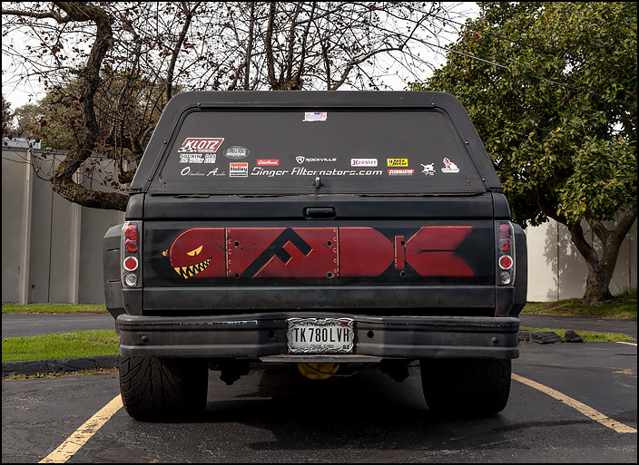 An old pickup truck with an F-Bomb painted on the tailgate. It is a bomb with a face on the front end and the letter F painted on the side.