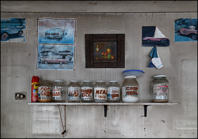 Jars with hand-painted labels sit next to a can of Raid insecticide on a shelf in an abandoned house. The wall around the shelf is decorated with photos of classic cars taken from old calendars. The jars contain popcorn, cornmeal, brown rice, brown sugar, peas, and lentils. The label on one says it contains Gods Force and Source. A painting of fruit hangs above the shelf.