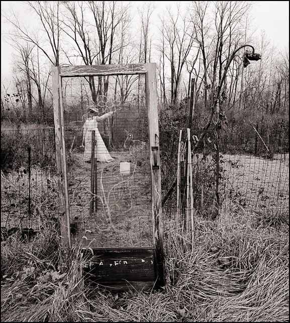 A weathered old wooden door used as a gate in the fence around one of the garden plots at the Fort Wayne Parks Department Community Garden. A scarecrow wearing a white dress is visible through the large window in the door.