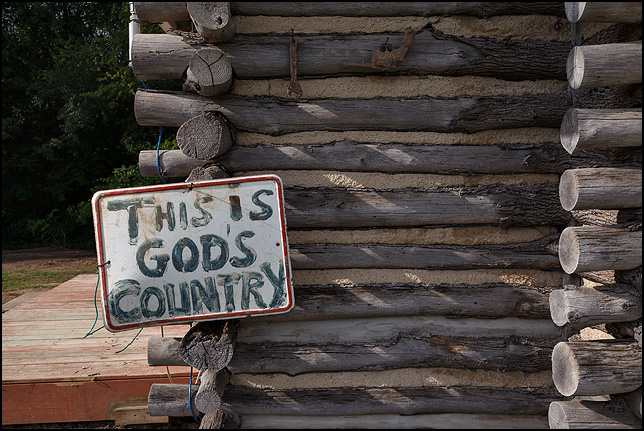 A hand-painted religious sign on the front of an old log cabin in rural Elkhart County, Indiana. This Is God's Country. A rusted handgun hangs above the sign.