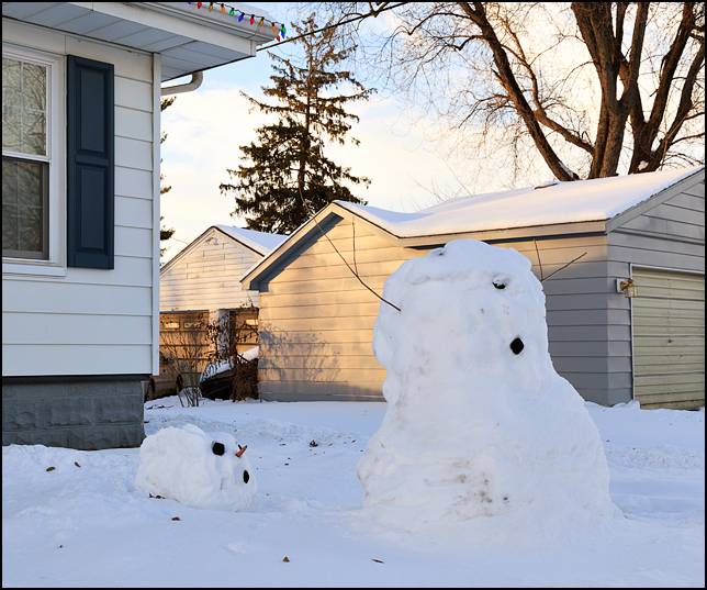 A headless snowman stands in front of a house on Clay Street in Mishawaka, Indiana. The head of the snowman is laying on the ground behind him, looking up at the top of the headless body.