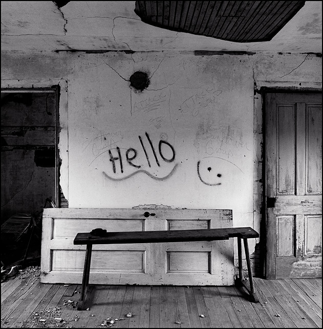 An old wooden bench and a weathered wood panel door sit in front of a wall covered in graffiti in a bedroom of an abandoned farmhouse in Indiana. The graffiti includes a spray-painted happy face and the word Hello.