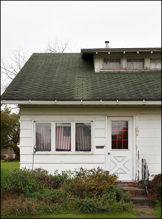 A small white house with an American flag in each of the three front windows. The house is on Antwerp Drive in the small town of Hicksville, Ohio.