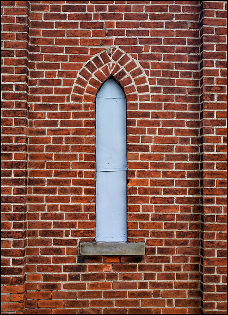 A boarded up pointed arch window on the steeple tower of an abandoned brick church at the corner of Hoagland Road and Clayton Road in southeast Allen County, Indiana. The window is covered in gray metal sheeting.