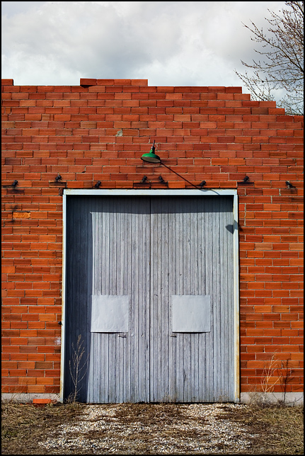 A green light with a large reflector hangs above a large garage door in an old abandoned brick church at the corner of Hoagland Road and Clayton Road in rural southeast Allen County, Indiana. The door is made of old blue painted wooden siding.