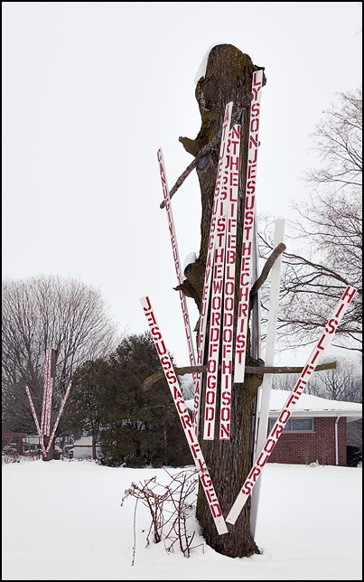 A dead tree with its branches cut off is covered in hand-painted religious signs about Jesus Christ.