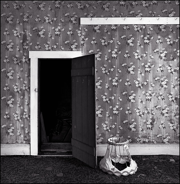 A little attic door in the wall of the bedroom in an abandoned farmhouse. An old torn-up lampshade sits next to the door in front of floral wallpaper.