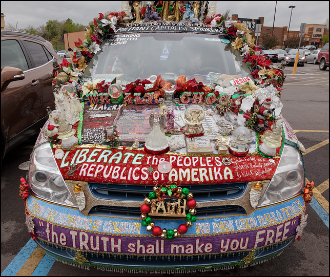 Crosses, angel figurines, snowglobes, Christmas decorations, and political signs cover the hood and bumper of a minivan.