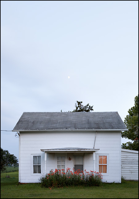 An old white farmhouse with a bed of tiger lilies in front of the porch, photographed at dusk with the moon rising in the sky above. County Road 900N in Huntington County, Indiana.