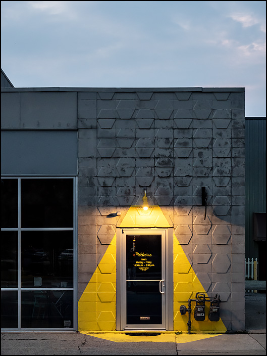 The front entrance of Lindis Restaurant on Main Street in downtown Fort Wayne, Indiana. The gray cinderblock building has the light projected from a bulb over the door painted on the front of the building in yellow paint.