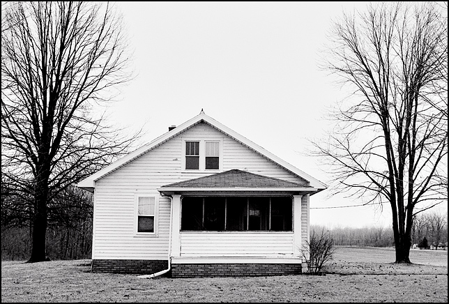 An abandoned house on Lower Huntington Road in the Waynedale area of Fort Wayne, Indiana. The house is a white woodframe bungalow surrounded by trees in front of an empty field.