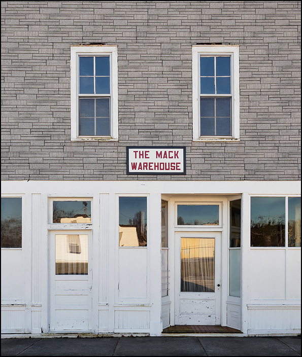 The Mack Warehouse, an old vacant building on 2nd Street in the small town of Wanatah, Indiana. The two story commercial building has asphalt shingle siding on the second floor and a white wooden storefront on the first floor.