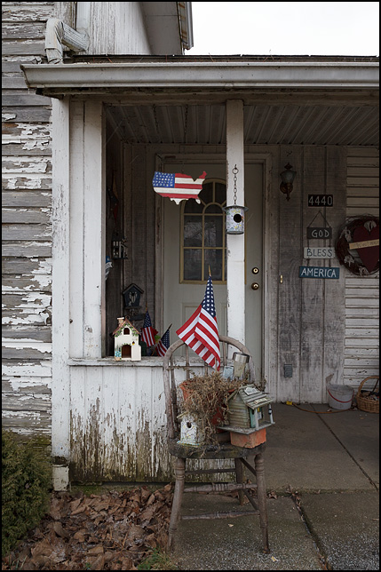 American flags and patriotic signs decorate the front porch of an old house with peeling paint on US-33 in the small town of Merriam, Indiana. Wooden cut-out of the United States painted like the American flag. God Bless America. Bentwood chair with several birdhouses sitting on it.