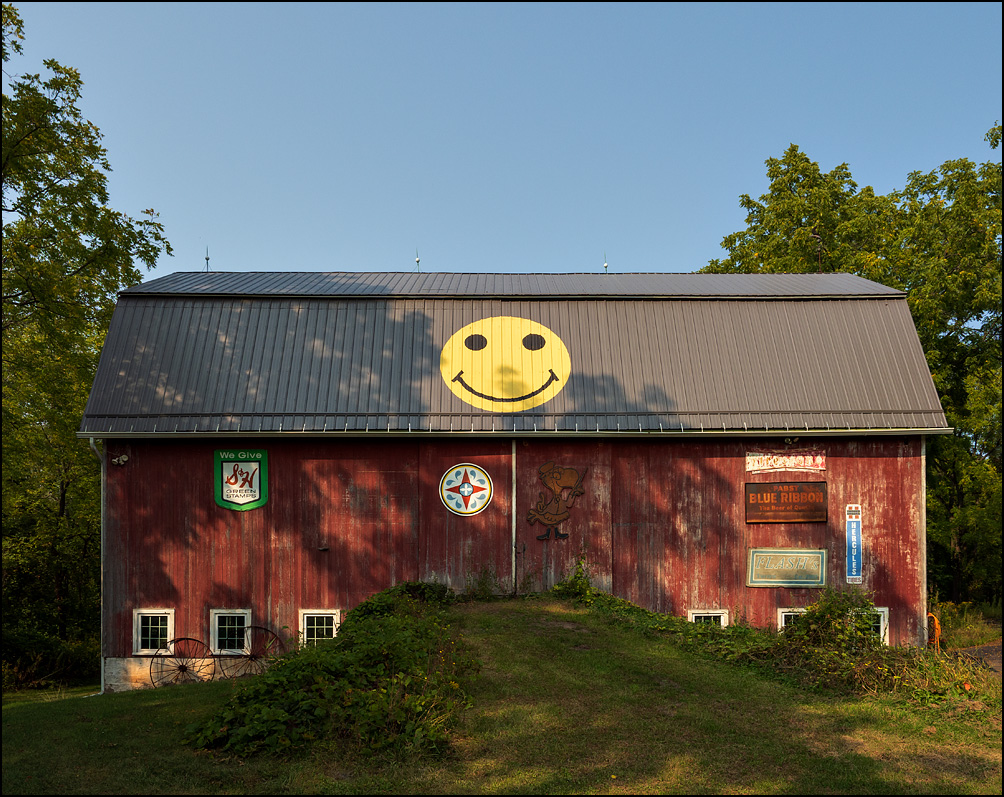 A red barn with a big yellow smiley face painted on the roof. State Road 31 in rural Monroe County, New York. There is a hex sign on the door and several old advertisements on the barn for Pabst Blue Ribbon beer, S&H Green Stamps, and Hercules tires.
