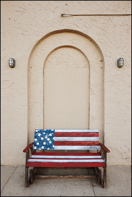 An old wooden glider bench painted like the American flag in front of a boarded-up stucco arched door on Broadway in Fort Wayne, Indiana. The bench stands in front of Little Shop of Lauras, a secondhand shop.