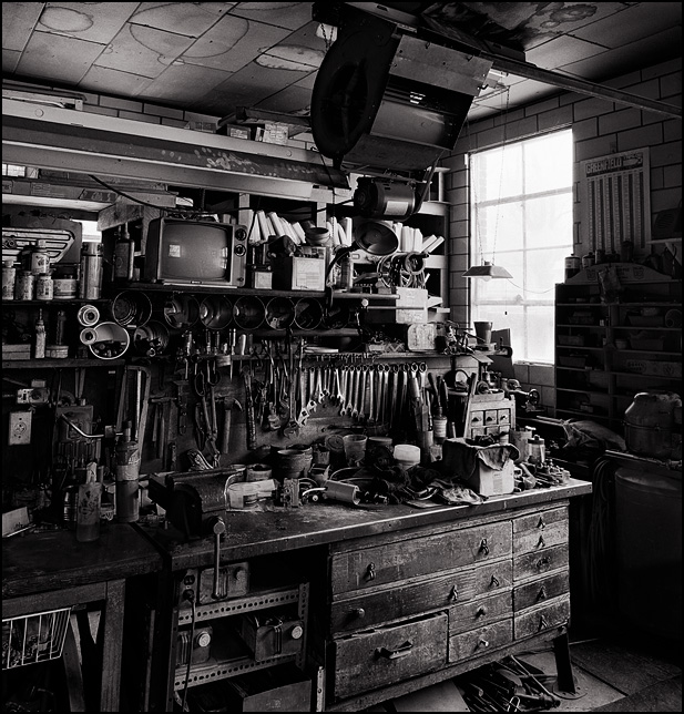 Wrenches and tools hang behind the cluttered old weathered wood workbench in the back room workshop at Waynedale Plumbing Supply. An ancient television sits on a shelf above the bench in the dimly lit room.