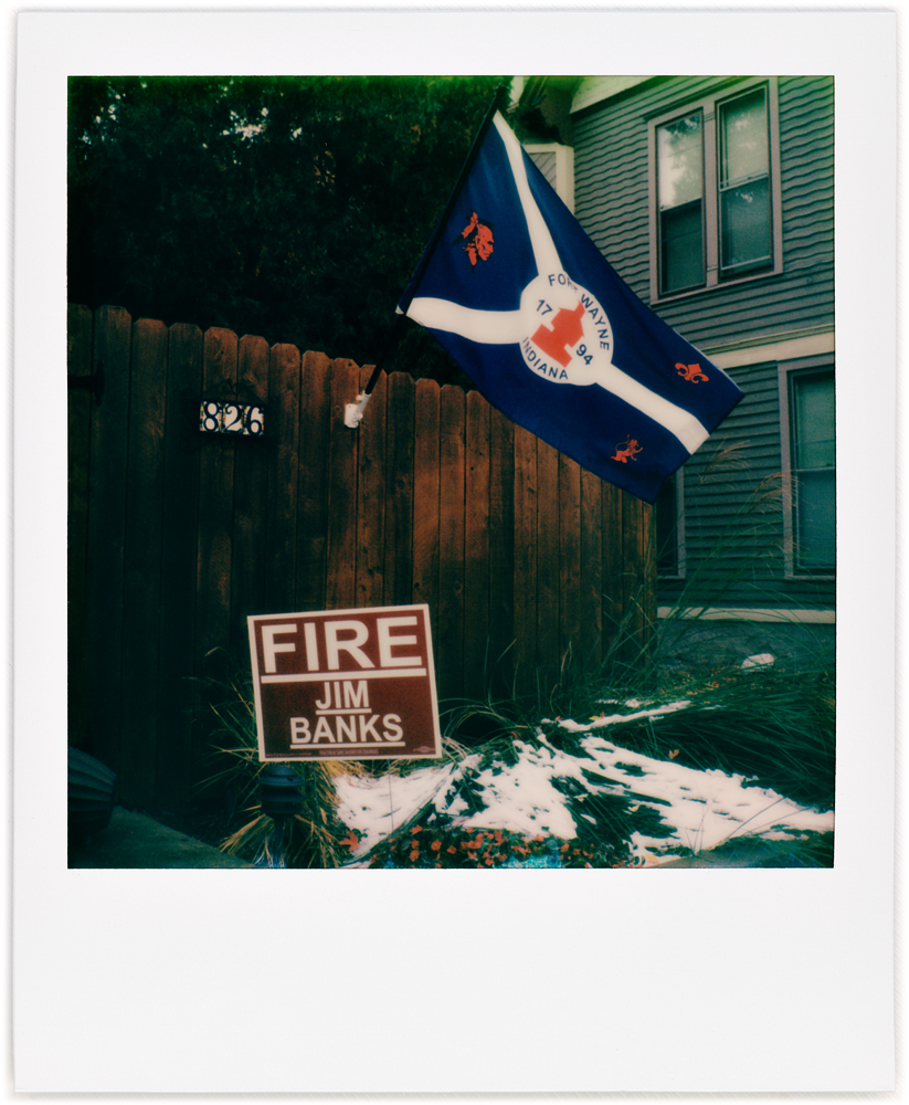 Polaroid snapshot of a wooden fence with the flag of the city of Fort Wayne and a political sign that says Fire Congressman Jim Banks. West Central neighborhood in Fort Wayne, Indiana.