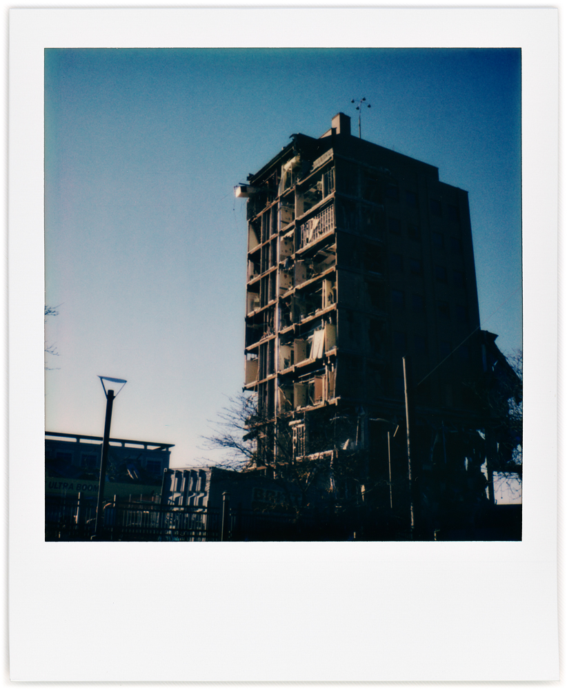 A Polaroid photo of the last remaining section of the partially demolished Saint Joseph Hospital in downtown Fort Wayne, Indiana. A brick tower with the steel frame and the rooms inside visible, silhouetted against the blue sky.