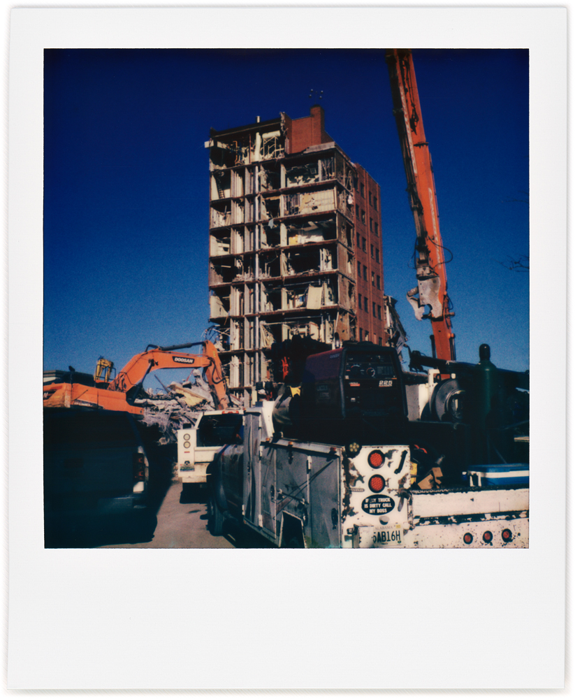 A Polaroid photo of demolition workers pickup trucks, excavators, and a crane parked in front of the partially demolished Saint Joseph Hospital in downtown Fort Wayne, Indiana.