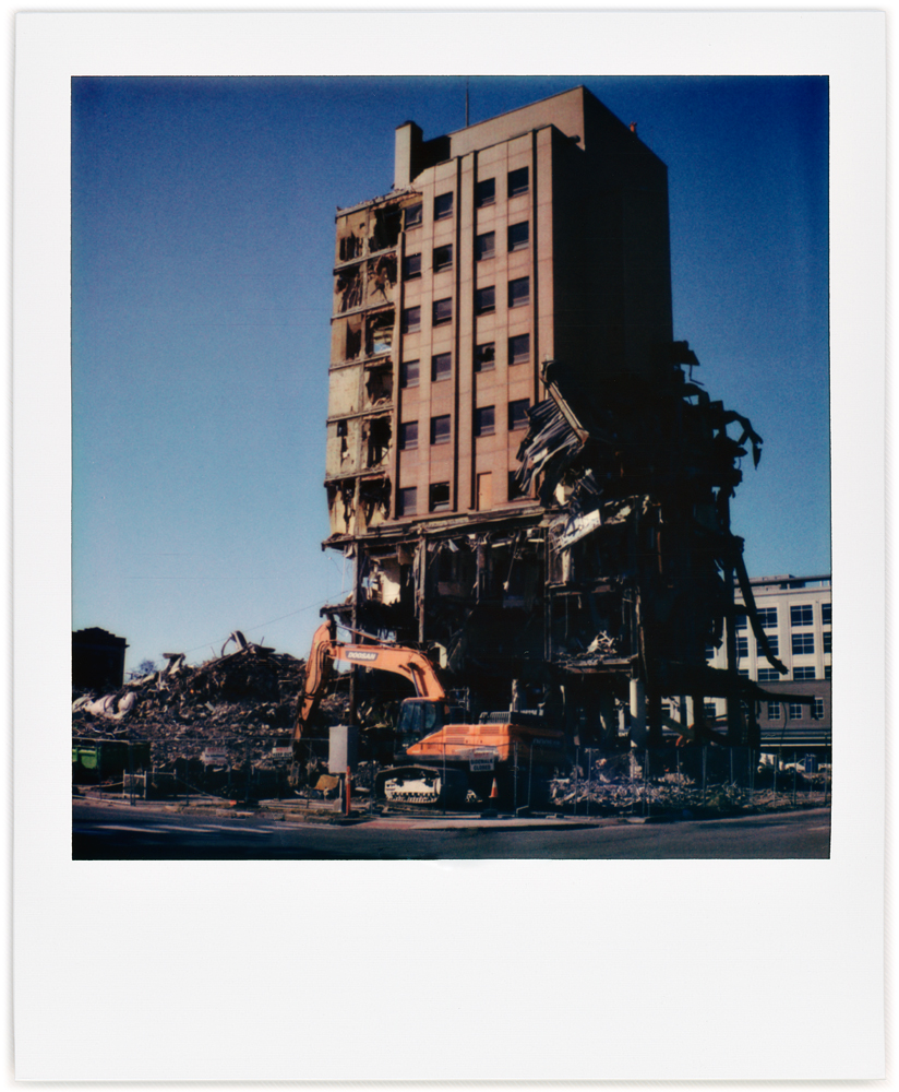 A Polaroid photo of the Saint Joseph Hospital demolition site in downtown Fort Wayne, Indiana. An excavator used to break apart the building stands in front of the partly demolished tower that used to be the north end of the building.
