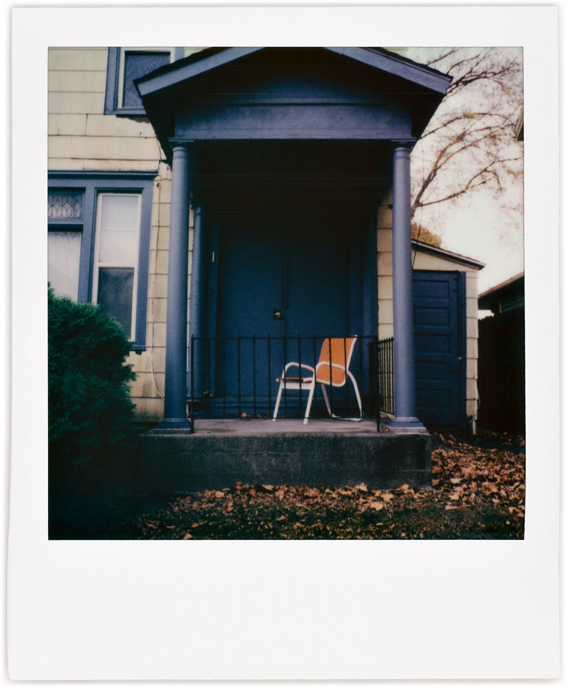 Polaroid snapshot of an old house with a blue door and an orange chair on the front step behind a wrought iron railing on Jefferson Boulevard in the West Central neighborhood in Fort Wayne, Indiana.