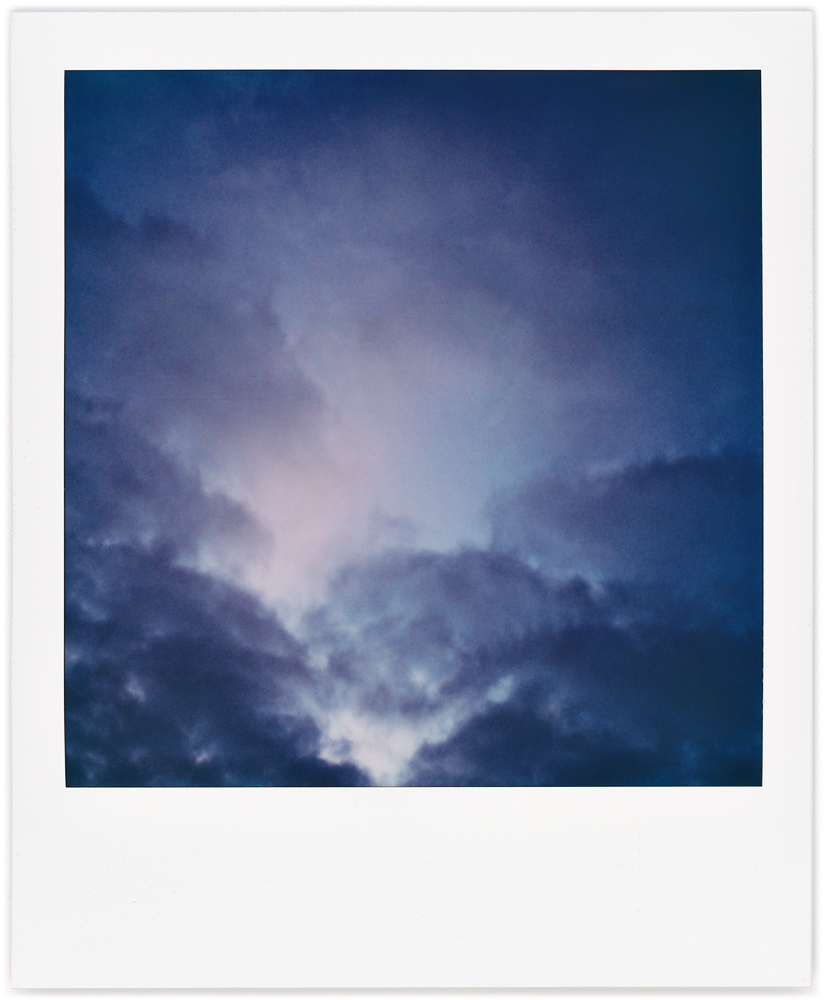 Polaroid abstract photograph of a V-shaped dark cloud formation with a round cloud above it in a field of bright sky.
