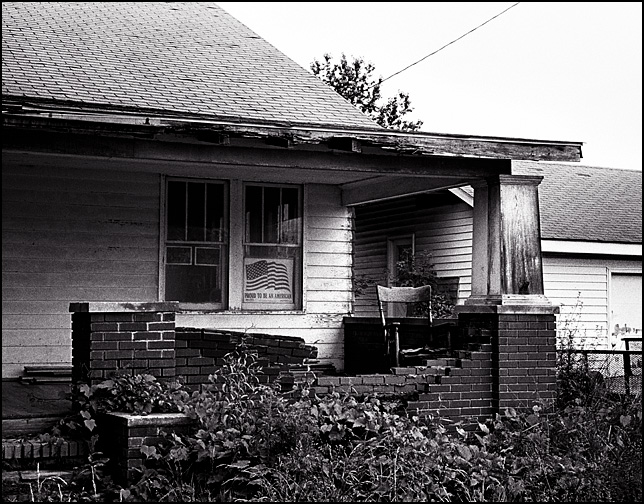 An old wooden rocking chair sits on the front porch of an abandoned house that has an American Flag sign in the window that says "Proud to be an American."