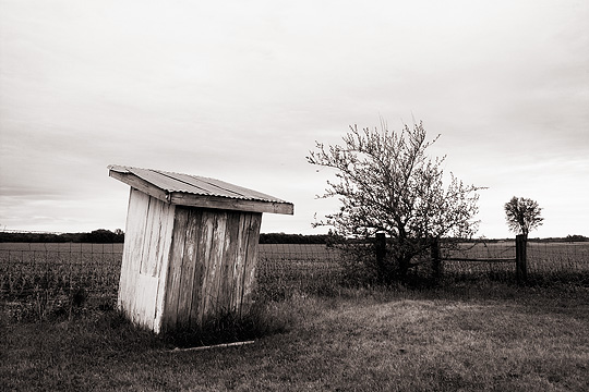 An outhouse behind the Lawton Christian Church in rural Pulaski County, Indiana. The little wooden outhouse faces toward a cornfield and a wire fence.