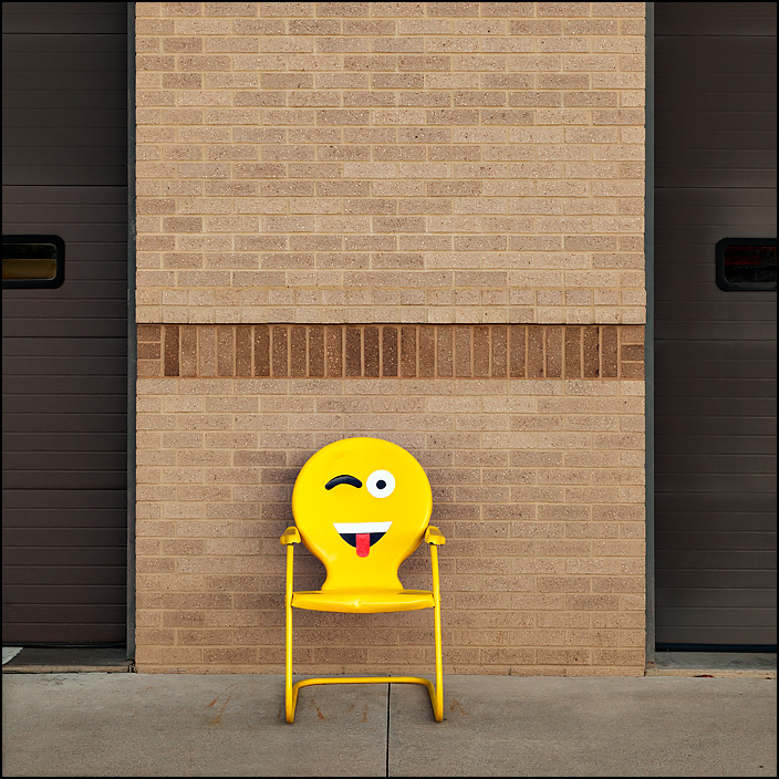 A yellow metal motel chair with a silly face painted on the back rest sits in front of Fire Station 10 at the corner of Crescent and Anthony in Fort Wayne, Indiana. The face on the chair is an Emoji winking and sticking out its tongue.