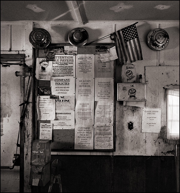 An American flag and a bulletin board on the wall of the office of Smith and Buckland Auto Parts, a junkyard in Fort Wayne, Indiana. The board is covered in signs spelling out the company's sales policies, but it also includes a political sign that says Indiana governor Mitch Daniels is a tax and spender who is targeting taxpayers for their money.