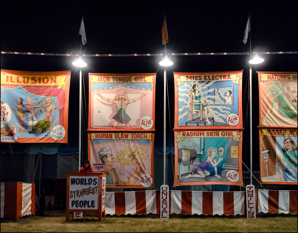 The entrance to a sideshow act called Worlds Strangest People at the 2018 Three Rivers Festival carnival in Fort Wayne, Indiana. The lonely carny sits at night in front of brilliantly illuminated banners advertising the circus freaks inside the tent.