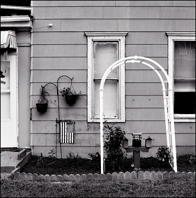 A small American flag hangs from a Welcome sign under a couple of hanging flower pots next to a wooden wedding arch in front of an old house on Taylor Street in Fort Wayne, Indiana. There are two small birdhouses under the arch.