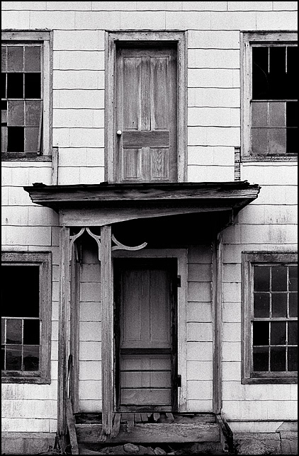The front of an abandoned farmhouse on US-33 in Noble County, Indiana. The porch has victorian gingerbread and there is a weathered wood door on the second floor opening to the roof of the porch.