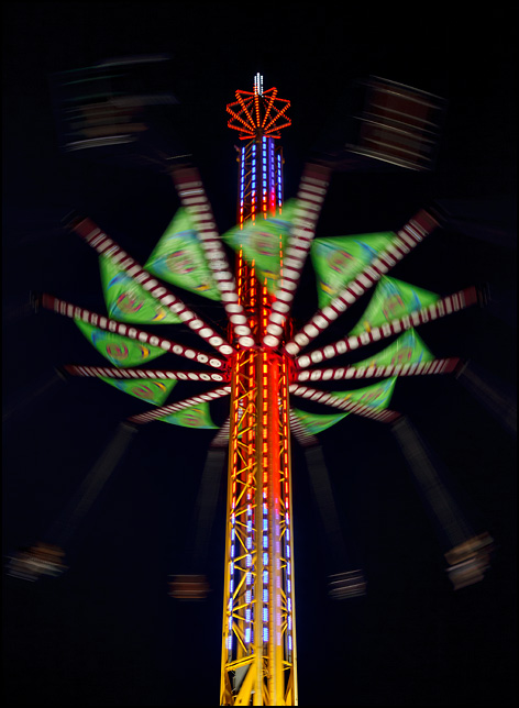 The Vertigo ride spins around the middle a tall tower at the 2016 Elkhart County Fair in Goshen, Indiana. The riders are a bright blur circling the tower in the night sky.