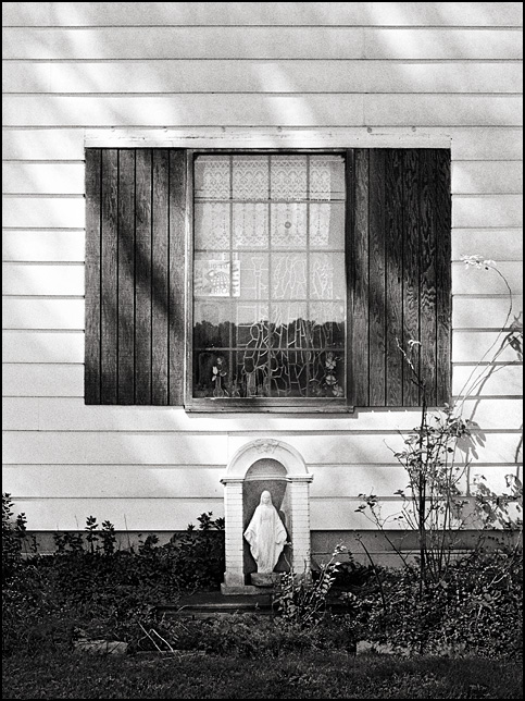 A statue of the Virgin Mary stands in front of Grover Schinbeckler's house in Dunfee, Indiana on the Whitley County side of County Line Road. A stained glass religious image hangs in the window above the statue of Saint Mary.