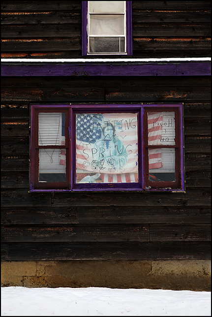 An old house in Fort Wayne that has an American Flag with a Native American on it hanging in the front window. Someone has drawn flowers and the Sun on the window and written, Waiting For Spring - Happy Days, on the window.