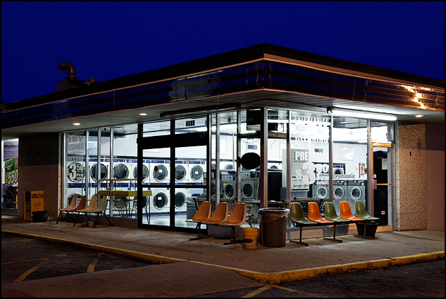The Wash House Laundromat glows with light in the early morning darkness. Plastic seats like those in airport waiting areas sit in front of the store's windows.