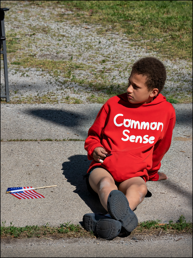 An adolescent African-American boy wearing a red hoodie that says, Common Sense.". He is watching the 2023 Waynedale Memorial Day Parade in the Waynedale area of Fort Wayne, Indiana.