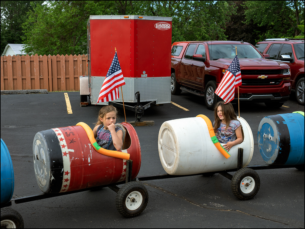 Two little girls sit in a train made of plastic barrels decorated with American flags in the staging area before the start of the 2023 Waynedale Memorial Day Parade in the Waynedale area of Fort Wayne, Indiana.
